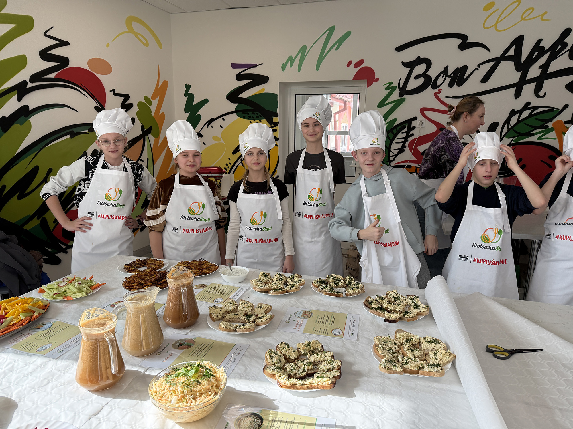 Dzieci w strojach kucharskich prezentują przygotowane posiłki. Na stole widoczne są kanapki z pastą jajeczną, surówka Colesław, placki warzywne, świeże warzywa, koktajle w dzbankach.